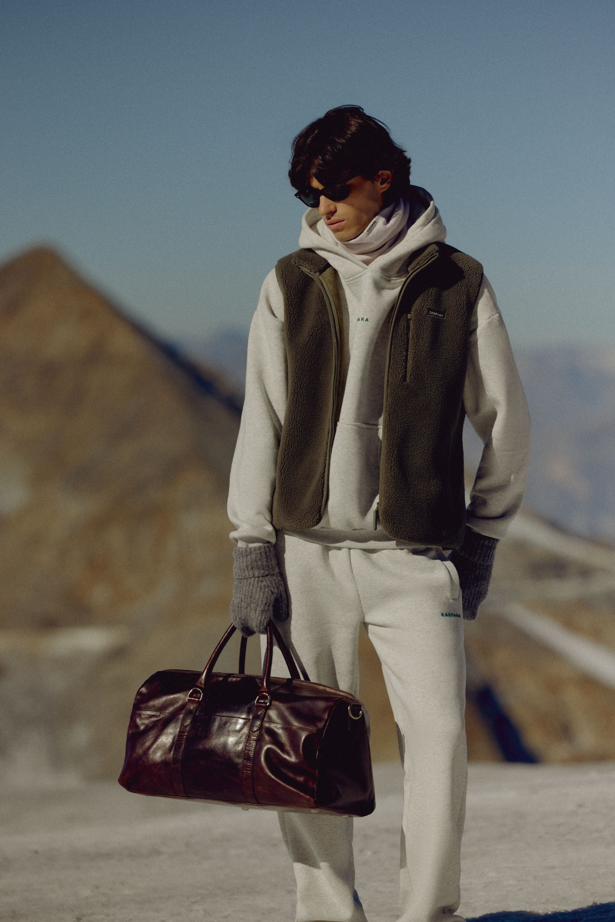 Man wearing a khaki CASPARA Cloudfleece vest holding a duffle bag in the mountains.
